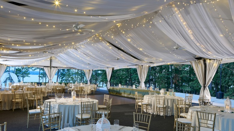 An elegant outdoor event space decorated with string lights and draped fabric, set with round tables and golden chairs for a celebration.