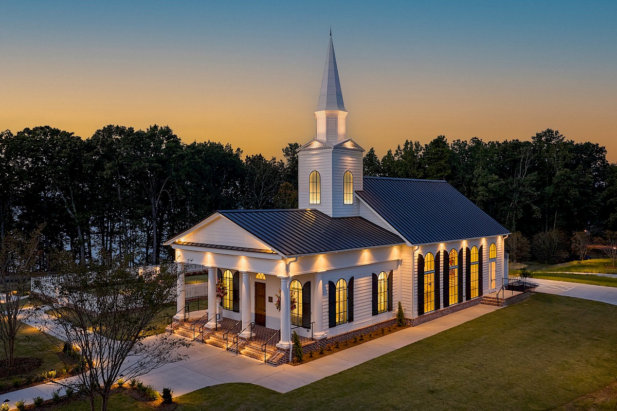 A church with white walls, tall windows, a steeple, surrounded by trees and a well-lit entrance at dusk.