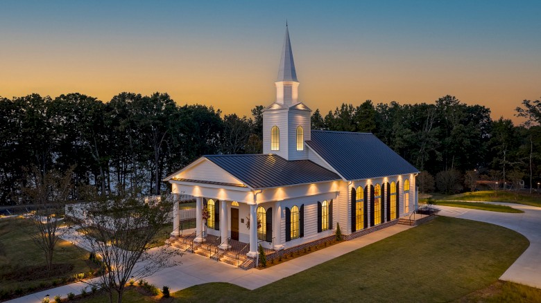 A church with white walls, tall windows, a steeple, surrounded by trees and a well-lit entrance at dusk.