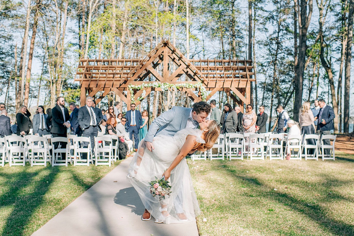 A couple shares a romantic kiss at an outdoor wedding ceremony surrounded by family and friends in a wooded area.