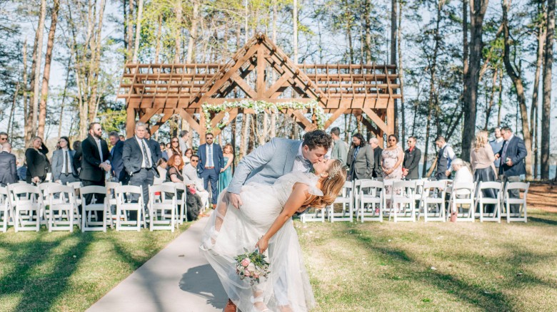 A couple shares a romantic kiss at an outdoor wedding ceremony surrounded by family and friends in a wooded area.