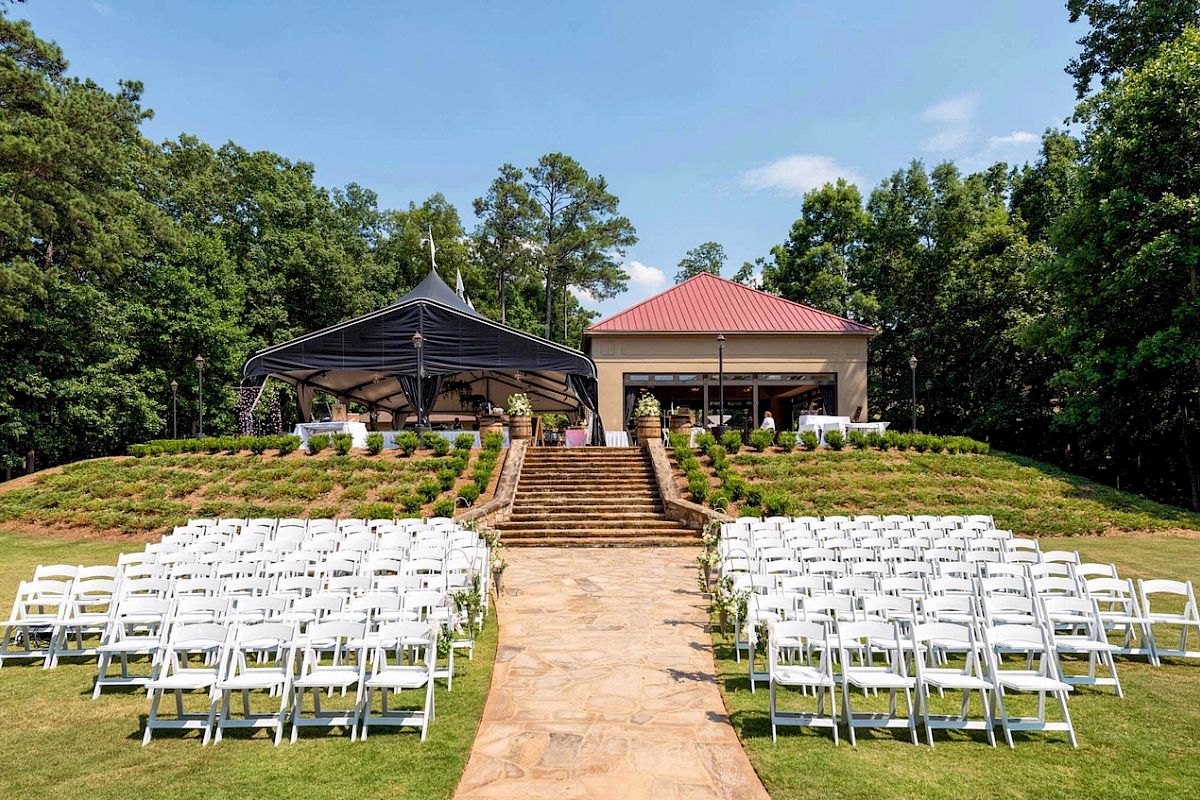 An outdoor wedding setup with rows of white chairs facing a staircase leading to a small building and a shaded area, surrounded by greenery.