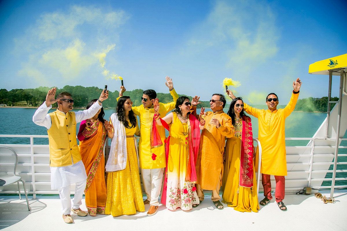A group of people dressed in colorful traditional attire celebrate with yellow smoke on a boat by the water under a sunny sky.