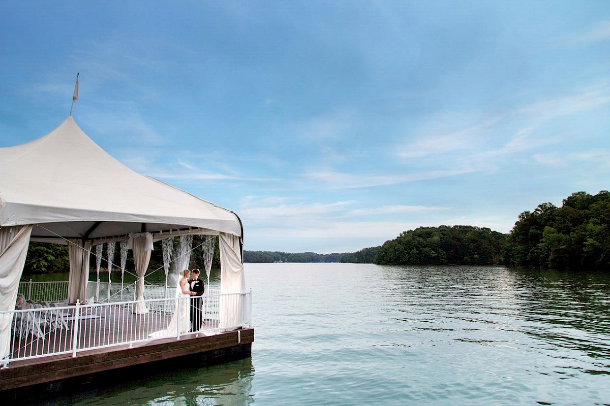 A couple stands under a white canopy on a pier by a calm lake with trees in the background, beneath a partly cloudy sky.