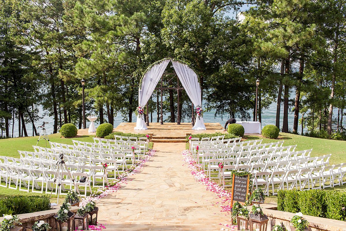 An outdoor wedding setup by a lakeside, featuring rows of white chairs, a decorated arch, and a stone path lined with flower petals, in a forested area.