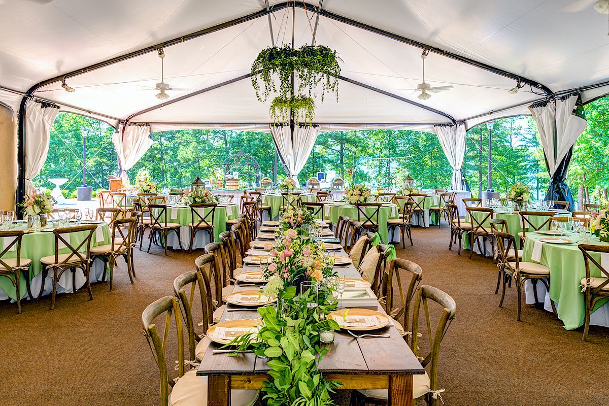 The image shows a beautifully decorated event tent with elegant tables and chairs, featuring a long centerpiece and greenery, set against a backdrop of trees.