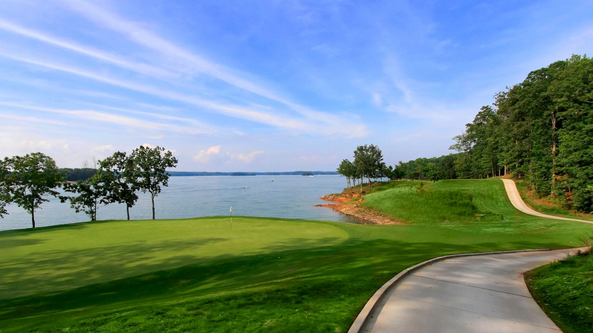 A scenic golf course by a calm lake, with a green, a paved path, trees lining the shore, and a blue sky above.