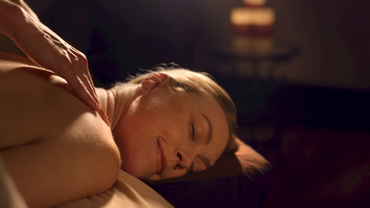 A person lying face down receiving a back massage in a dimly lit room, eyes closed and relaxed, peaceful mood, soft warm light.