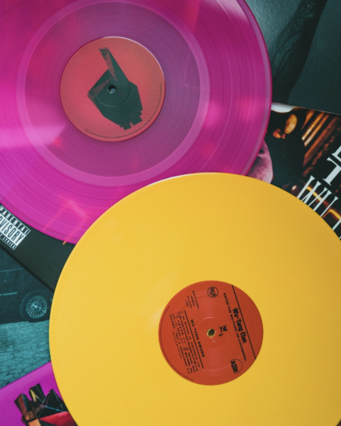 Two colored vinyl records, one pink and one yellow, lie atop album covers, featuring various designs and text.