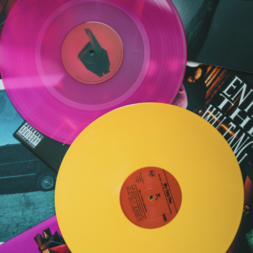 Two colored vinyl records, one pink and one yellow, lie atop album covers, featuring various designs and text.
