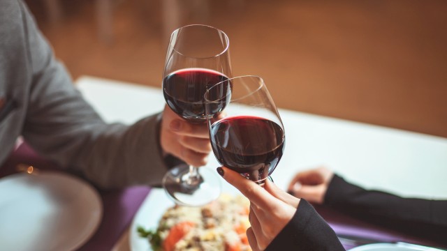 Two people clink glasses of red wine over a table setting with plates and a dish in the background.