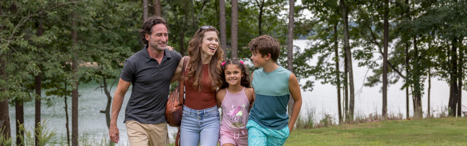 A happy family of four, including two kids, walking arm-in-arm through a grassy park near a lake, enjoying a joyful moment together.