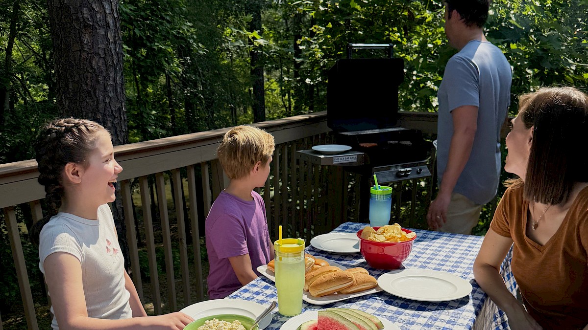 A family is having a barbecue dinner outdoors on a wooden deck surrounded by trees, with food like watermelon, chips, and drinks.