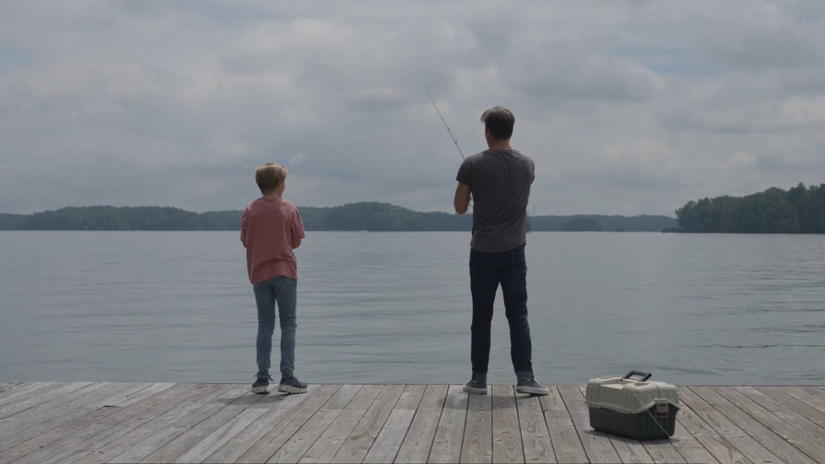 A man and a boy fish together on a dock by a calm lake with cloudy skies, and a tackle box sits nearby.