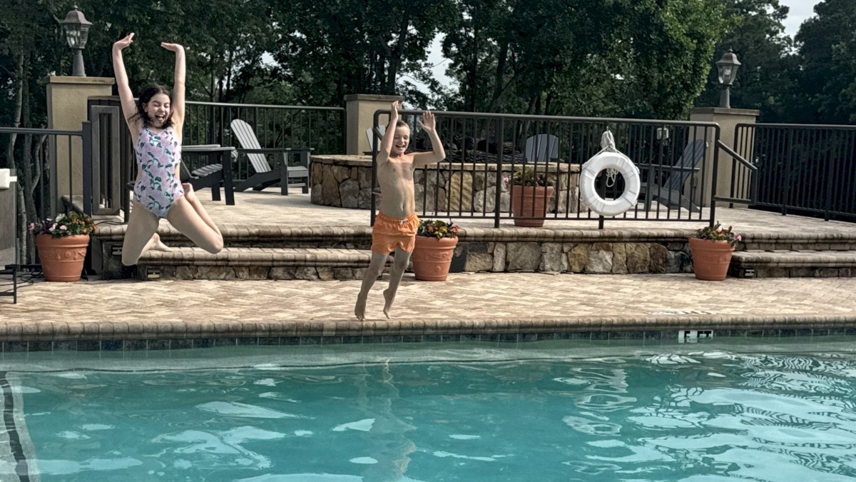 Two kids jump into a pool from the deck on a cloudy day.