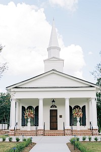 A white church with black-shuttered windows, flower arrangements, and a steeple against a partly cloudy sky.
