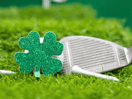 A golf club is lying on the grass with a sparkly green four-leaf clover decoration in front of it.