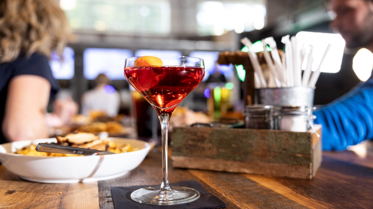 A red cocktail with an orange garnish on a wooden table, surrounded by food, straws, and people in a lively restaurant setting.