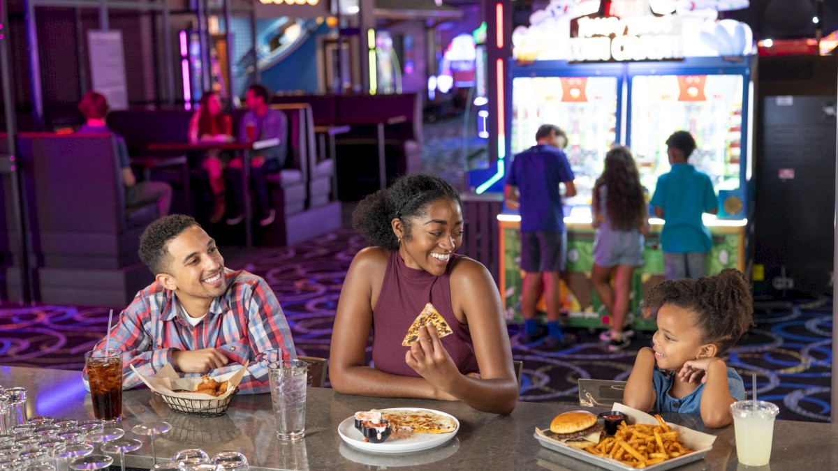A family enjoys pizza and fries at a lively arcade with bright lights and games in the background.