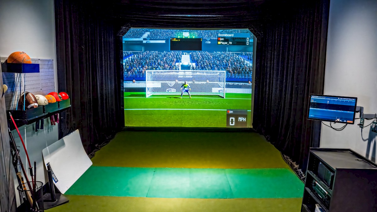 An indoor golf simulator room with a digital screen showing a soccer penalty shootout.