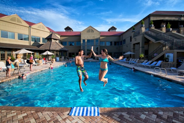 People enjoy swimming and jumping into a pool at a sunny hotel courtyard with lounge chairs and umbrellas.