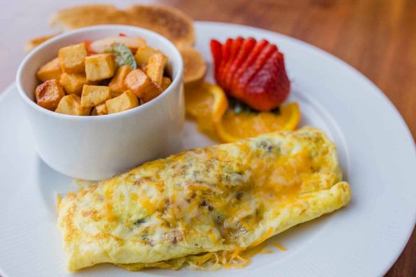 A plate with a cheesy omelette, a bowl of diced potatoes, sliced strawberries, orange slices, and toast in the background.