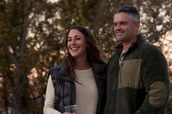 A smiling couple stands outdoors in front of trees, dressed in casual, warm clothing. The woman holds a clear cup in her right hand.