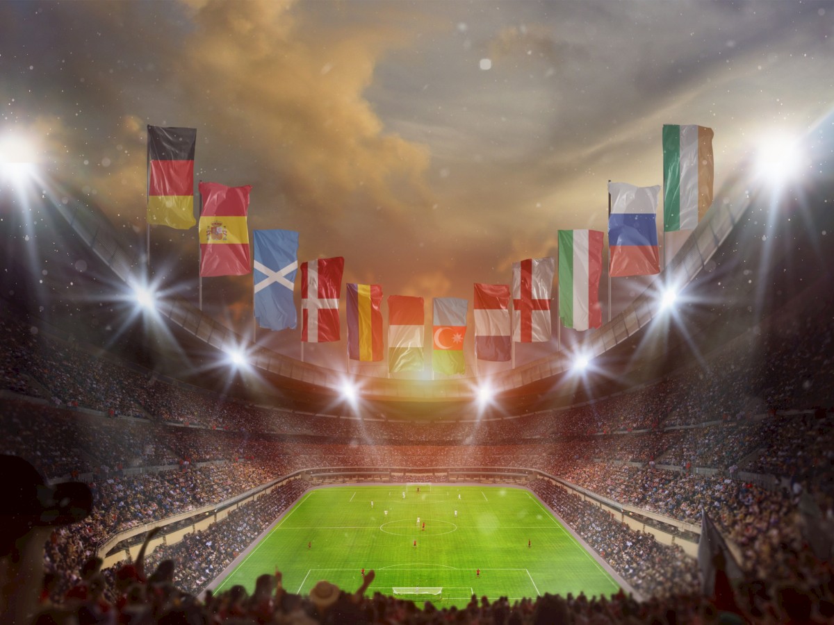This is an image of a packed stadium with football match underway under a dramatic sky, overhead flags from various countries fly in the breeze.