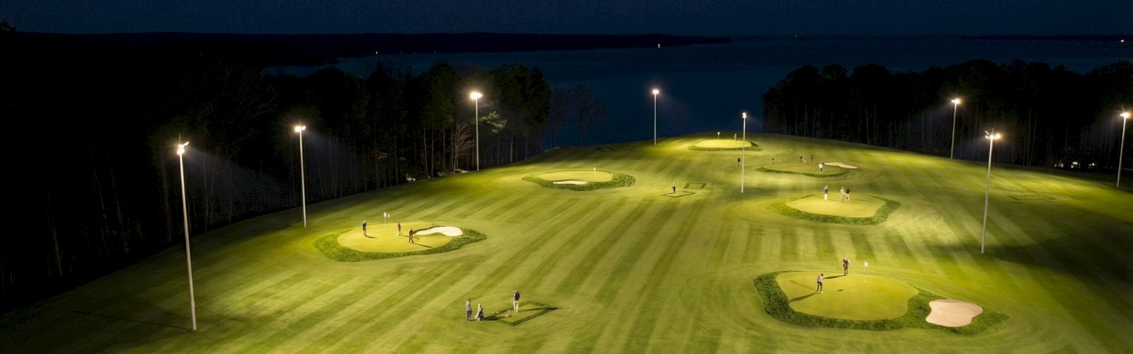 Nighttime on a golf course with players practicing putting under bright lights near a body of water.