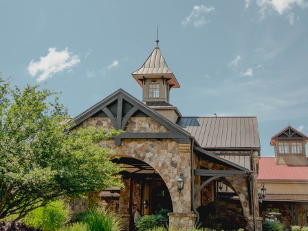 A charming stone building with a tower, dark roof, and lush landscaping under a partly cloudy sky.