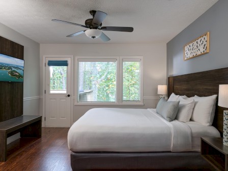 A cozy bedroom with a large bed, wooden furniture, a ceiling fan, wall art, and a window showing greenery outside.