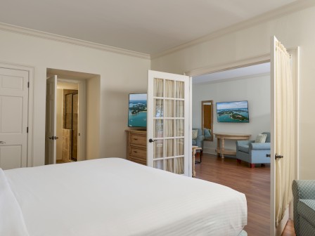 This image shows a bedroom with a view of a sitting area and TV through a French door, decorated in a light, neutral color palette.