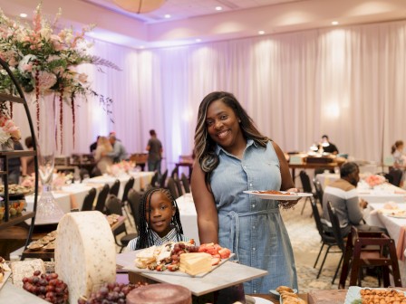 A woman and a girl are smiling at a buffet table with cheese, grapes, cake, and snacks in a decorated event hall.