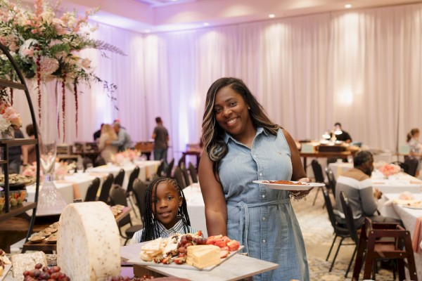 A woman and a girl are smiling at a buffet table with cheese, grapes, cake, and snacks in a decorated event hall.