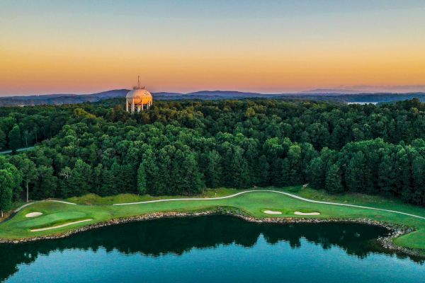 The image shows a picturesque landscape with a dome structure surrounded by dense trees, a golf course in the foreground, and a body of water.