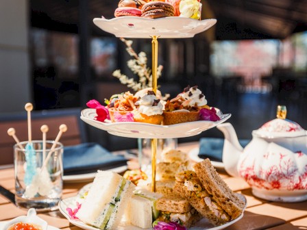 A three-tiered tray with assorted desserts, sandwiches, and pastries on a wooden table set for tea.