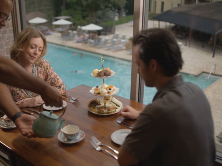 Two women and one man enjoy afternoon tea by a swimming pool with a tiered tray of desserts, while a server pours tea in a bright room.