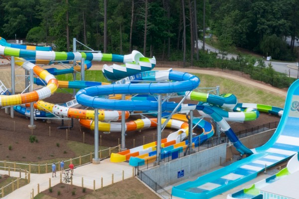 The image shows an outdoor water park with multiple colorful water slides and a ramp, surrounded by trees and pathways.