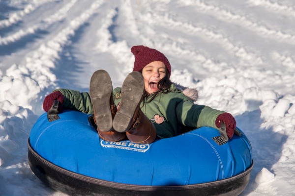 A person wearing a red hat and green jacket rides a blue snow tube down a snowy slope, smiling with legs up in the air.