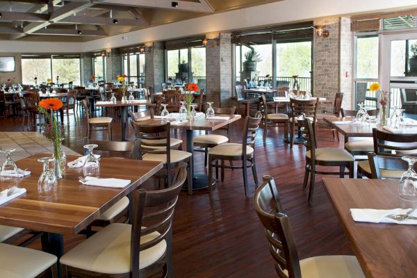 The image shows a spacious restaurant with wooden tables and chairs, set with napkins, glasses, and single-stem flowers in vases.