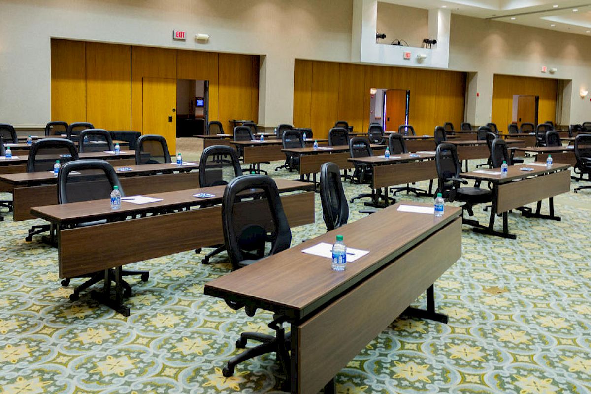 An empty conference room with long tables, office chairs, bottled water, and paper on the tables. The room has a carpeted floor and closed doors.