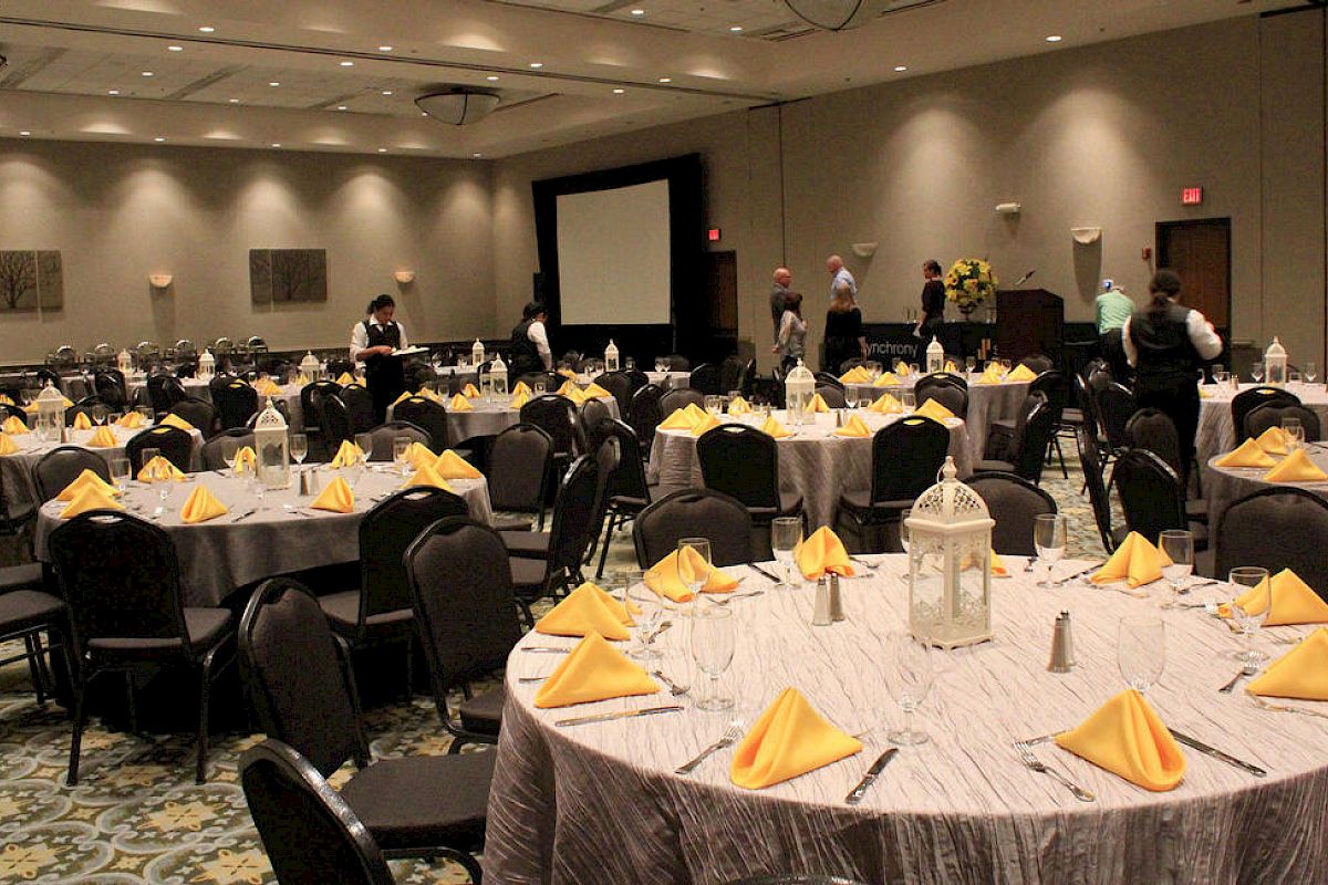 A banquet hall is set up with round tables covered with gray tablecloths and yellow napkins. Some people can be seen arranging items at the back.
