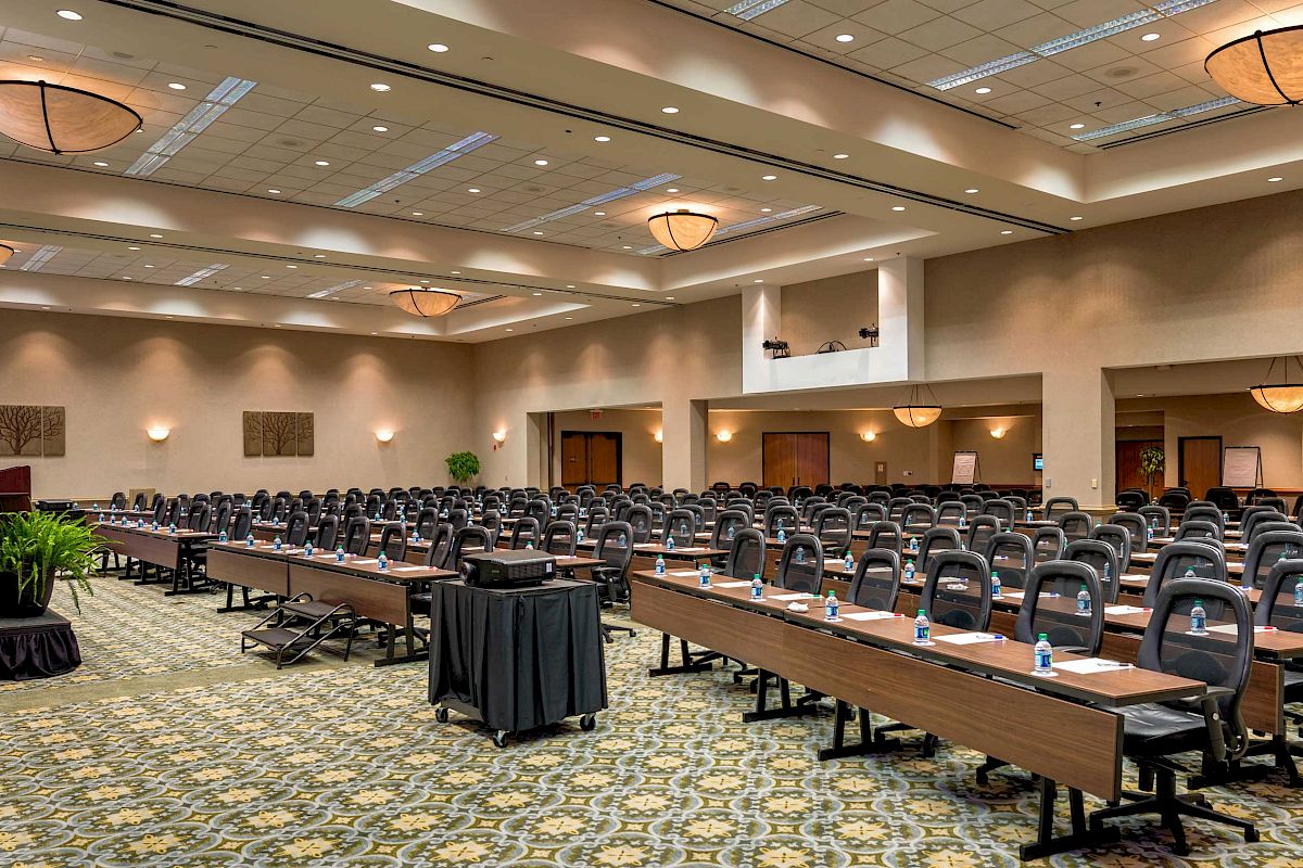 The image shows a large conference room set up with tables, chairs, and a podium on the left side. The room is well-lit with chandeliers.