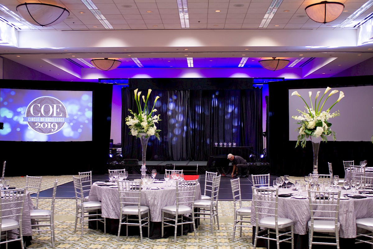 A fancy conference setup with round tables, white chairs, centerpieces, a stage with purple lighting, and projection screens displaying “COE 2019”.