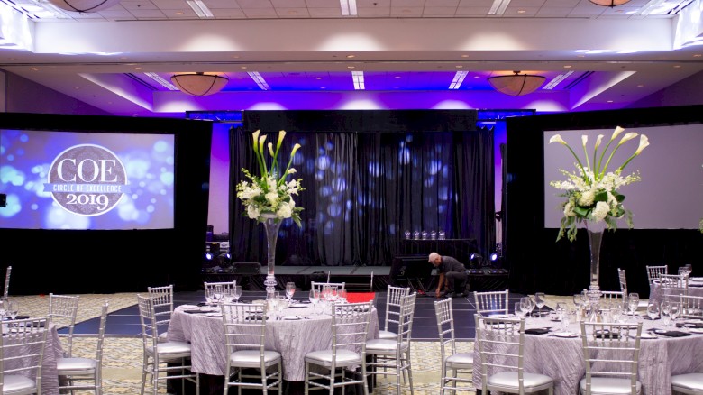 A fancy conference setup with round tables, white chairs, centerpieces, a stage with purple lighting, and projection screens displaying “COE 2019”.