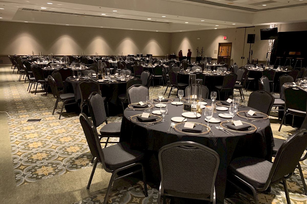 The image shows a large banquet hall set up for an event with round tables covered in black tablecloths, each arranged with place settings.