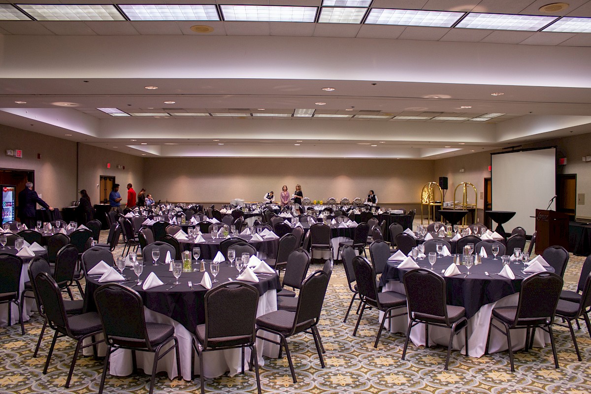 A banquet hall set for a formal event, with round tables dressed in white linens, black chairs, and neatly arranged place settings, ready for guests.