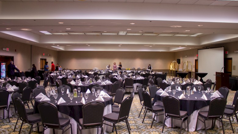 A banquet hall set for a formal event, with round tables dressed in white linens, black chairs, and neatly arranged place settings, ready for guests.