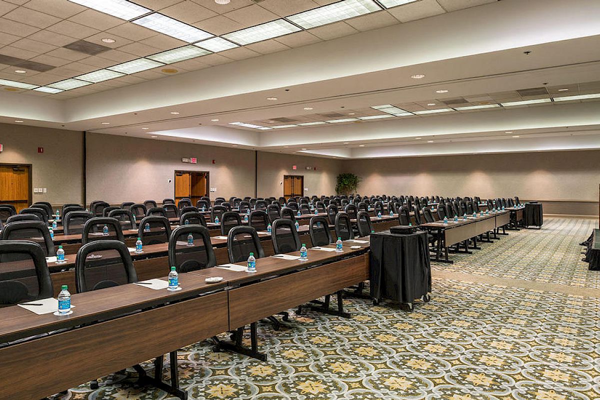 The image shows a large conference room with rows of tables and black chairs. Each table has bottled water and notepads. The room is empty.