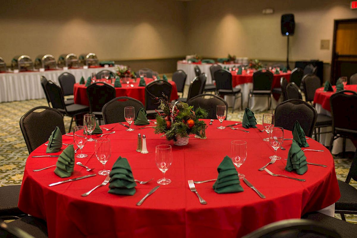 The image shows a banquet hall set up for a festive event with tables covered in red tablecloths and green napkins folded like Christmas trees.